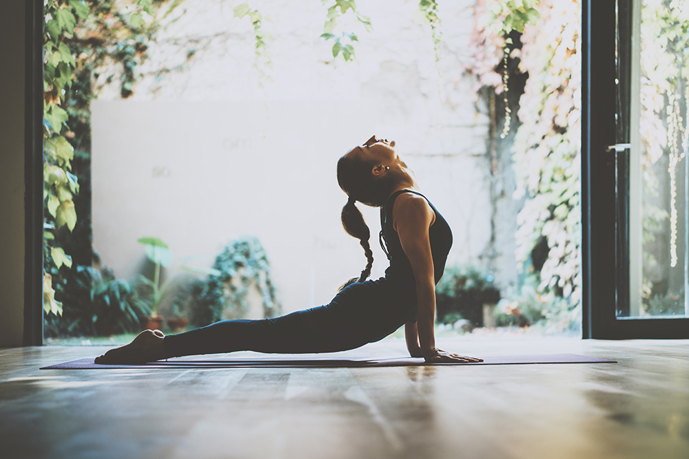 Síndrome de Burnout: Mulher branca praticando yoga. Aos fundos diversas plantas compõem o ambiente